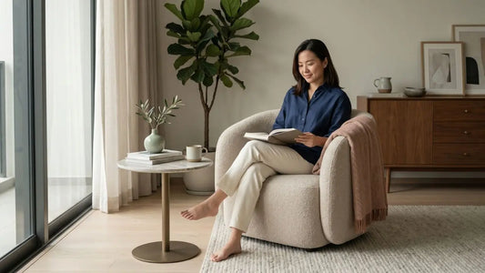 Woman reading in a rounded cream armchair beside a compact side table in a softly lit solo home living room.