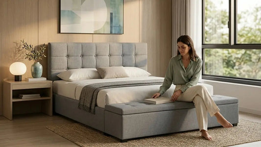 Woman sitting on an upholstered grey bed frame in a calm modern bedroom with a padded storage bench.